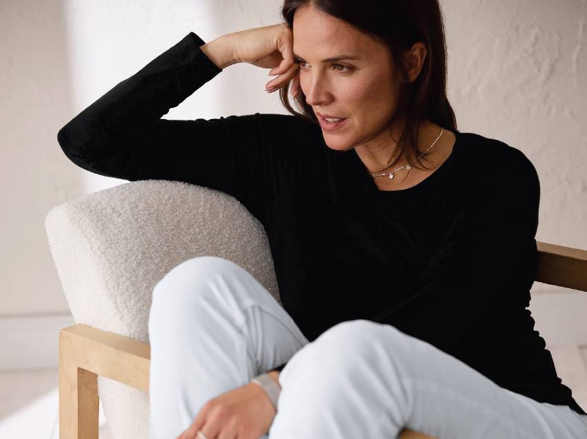 Жена в черна блуза и бели панталони, седнала на стол.