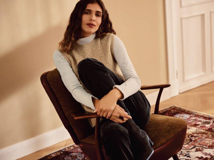 Жена в плетен елек и панталон, седнала на фотьойл в стая.