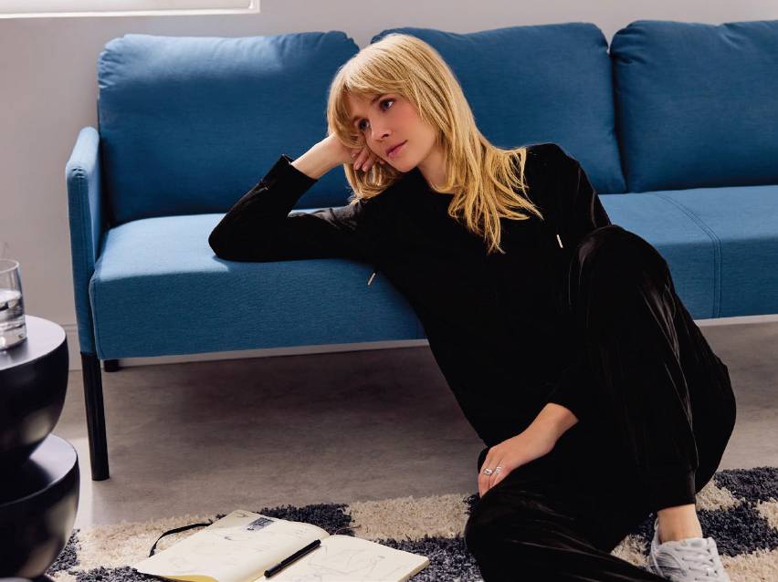 Жена седи на килим до син диван, скицирайки в тетрадка.