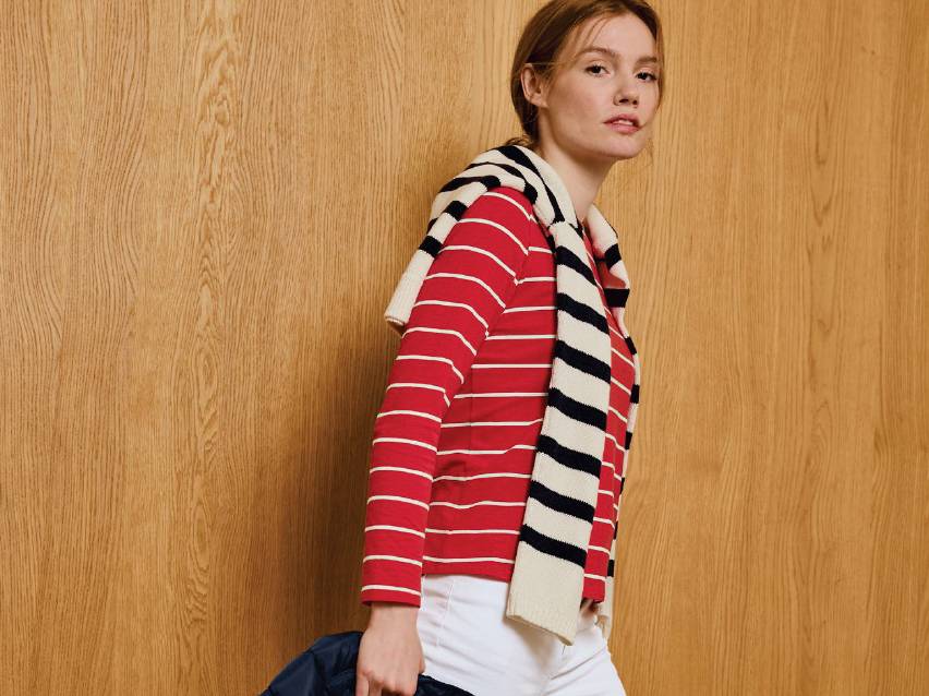 Жена в червена раирана блуза и бял панталон, с раиран пуловер на раменете.