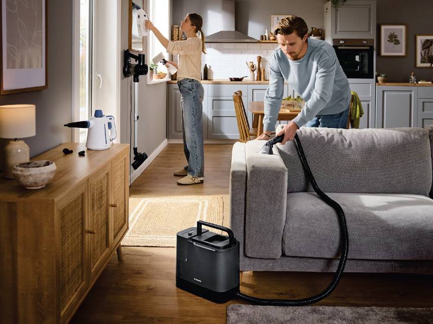 Мъж почиства диван с уред за почистване на тапицерия, жена почиства прозорец с парочистачка.