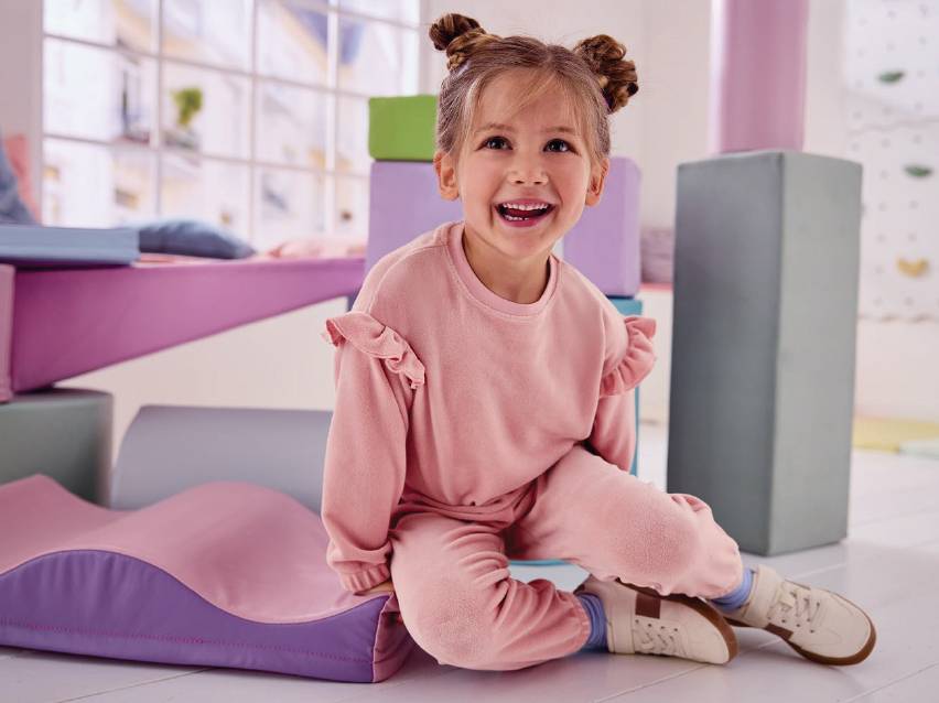 Усмихнато момиченце в розов суичър и панталон, седнало сред меки модули за игра.