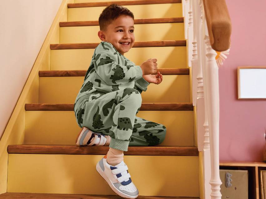 Момче в зелен комплект с динозаври седи на стълбите и се усмихва.