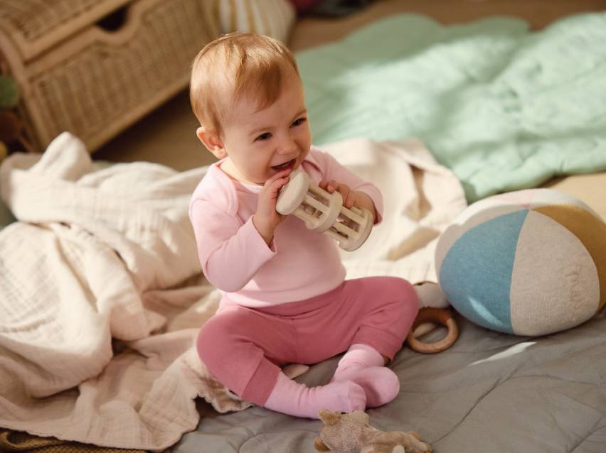 Бебе в розови дрехи, седнало на одеяло, играе с дървена дрънкалка.