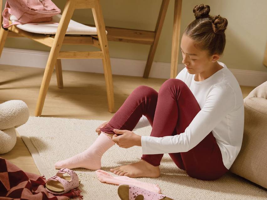 Момиче в червени клинове и бяла блуза обува розови чорапи на килим.