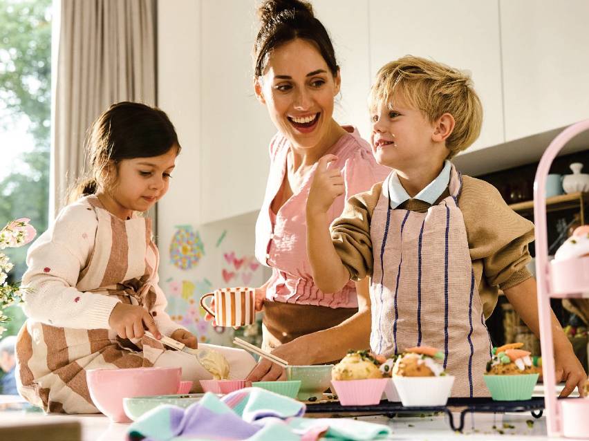Майка и две деца пекат кексчета в кухнята, използвайки купи и прибори.