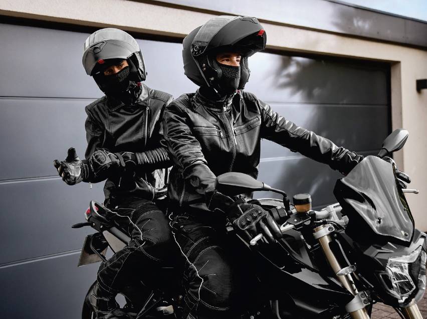 Двама мотористи с каски и кожени якета на мотоциклет пред гараж.