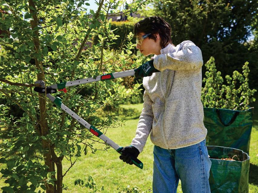 Жена подрязва храст с ножица за клони, облечена в сив суичър и предпазни очила.