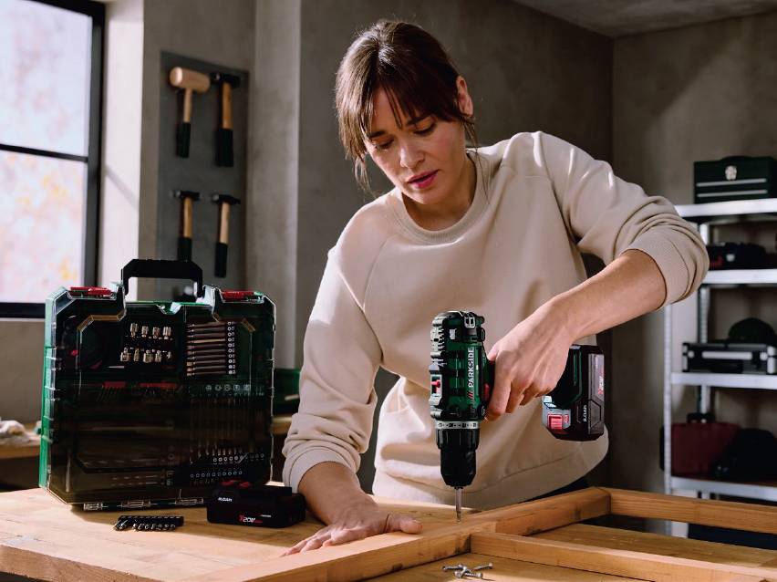 Жена използва акумулаторна бормашина, докато сглобява дървена рамка, до нея има кутия с битове.