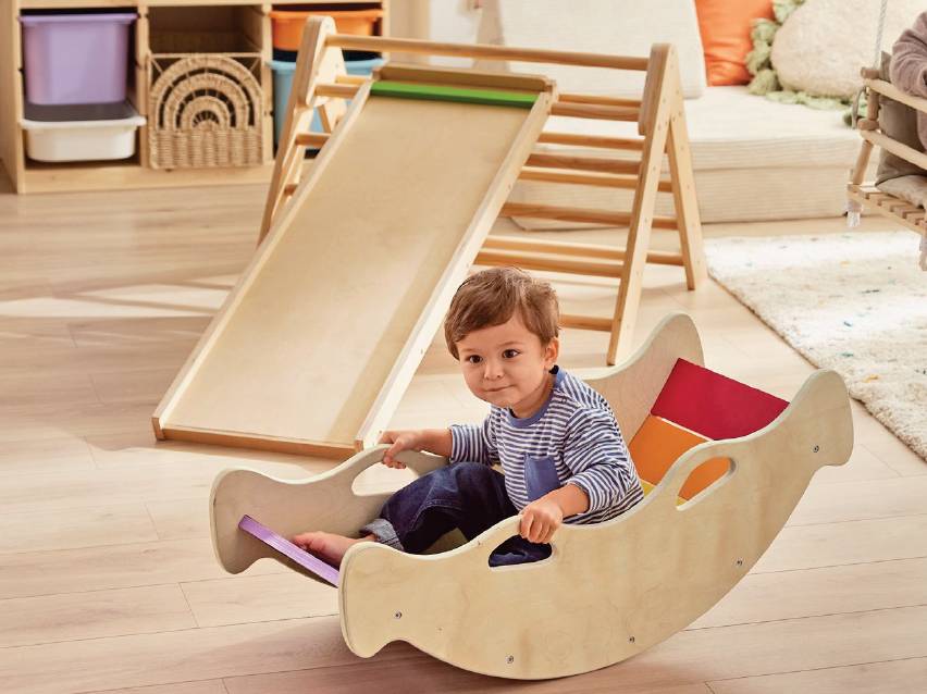 Дете в дървена люлка, до него дървена катерушка с пързалка в детска стая.