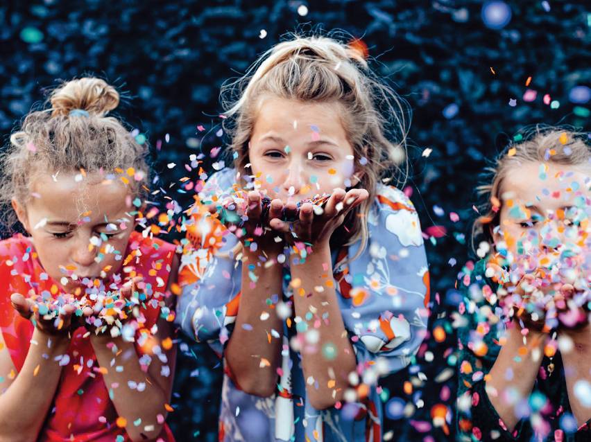 Три момичета духат конфети, празнувайки щастливо.
