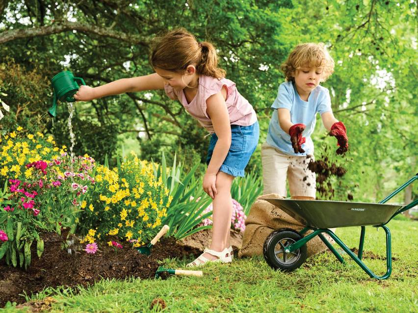 Две деца се забавляват в градината, поливайки цветя и пренасяйки пръст с градинска количка.