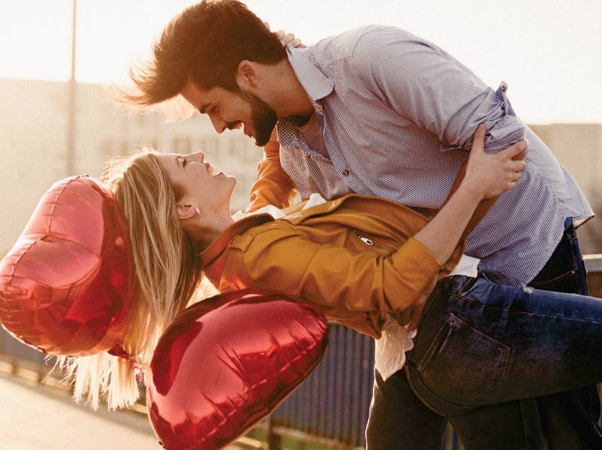 Влюбена двойка се смее, докато мъжът държи жената, а тя държи червени балони във формата на сърце.