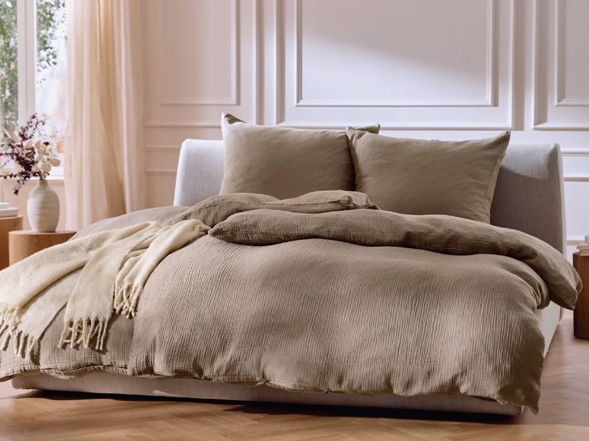 Спалня с бежово спално бельо от муселин, възглавници и одеяло.