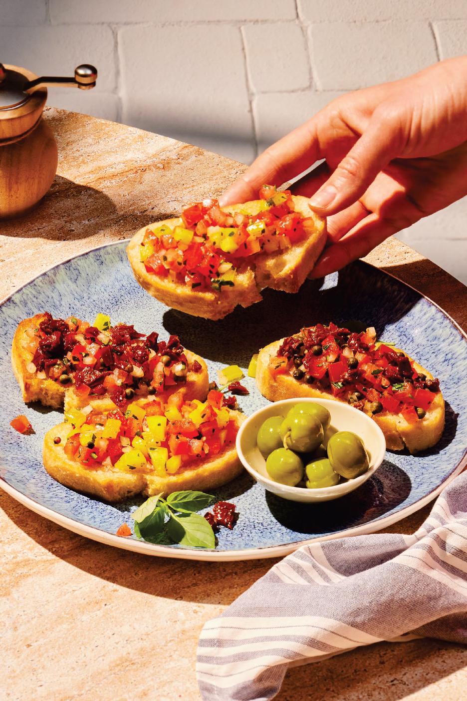 Ръка взима брускета с домати и чушки от синя чиния, до други брускети и маслини.