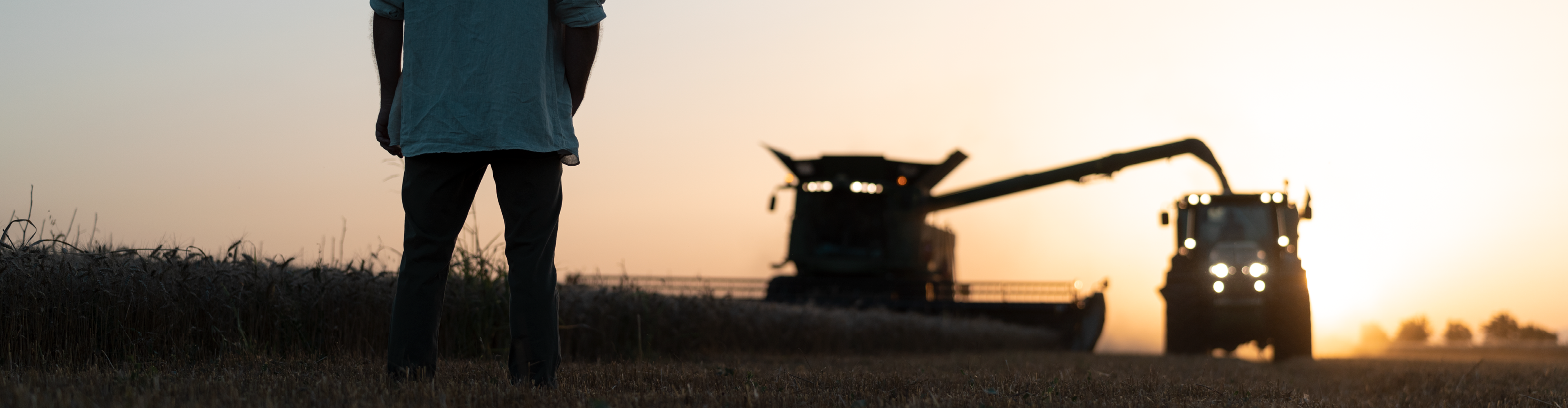 Фермер наблюдава комбайн по време на залез.
