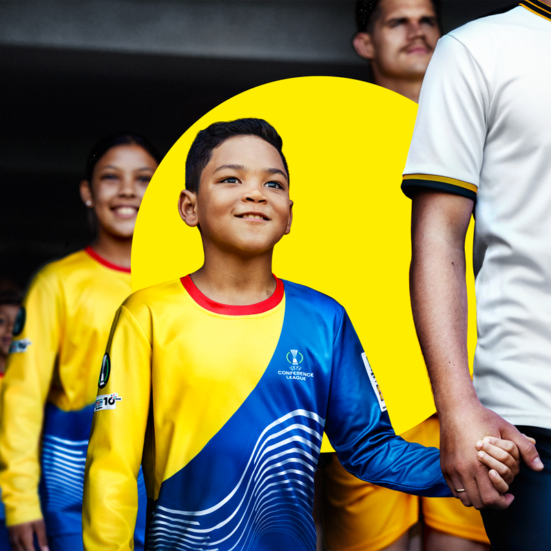 Младо момче в жълта и синя футболна фланелка държи ръката на възрастен.