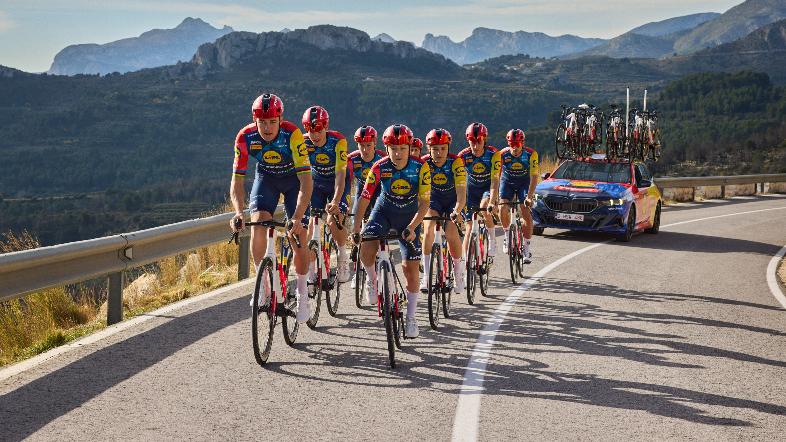 Колоездачи в екипи с логото на хранителен магазин, каращи по планински път, следвани от кола.