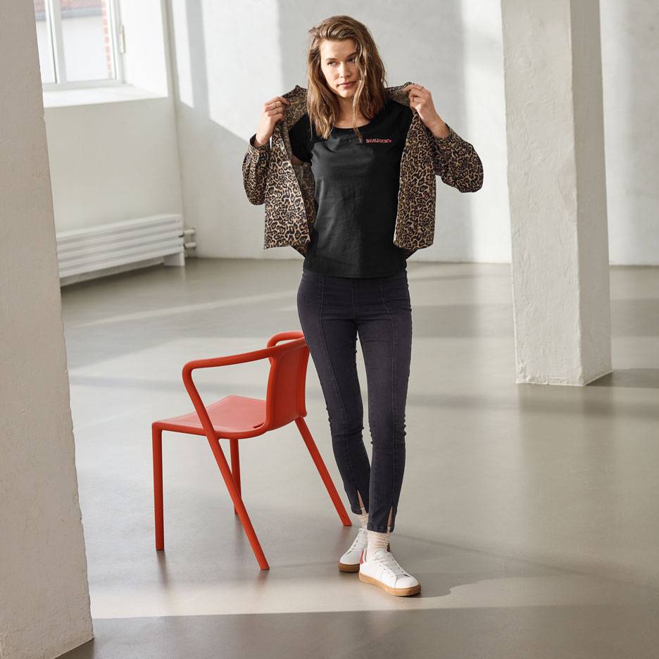 Жена в черна тениска с надпис, дънки и яке с леопардов принт, стояща до червен стол.