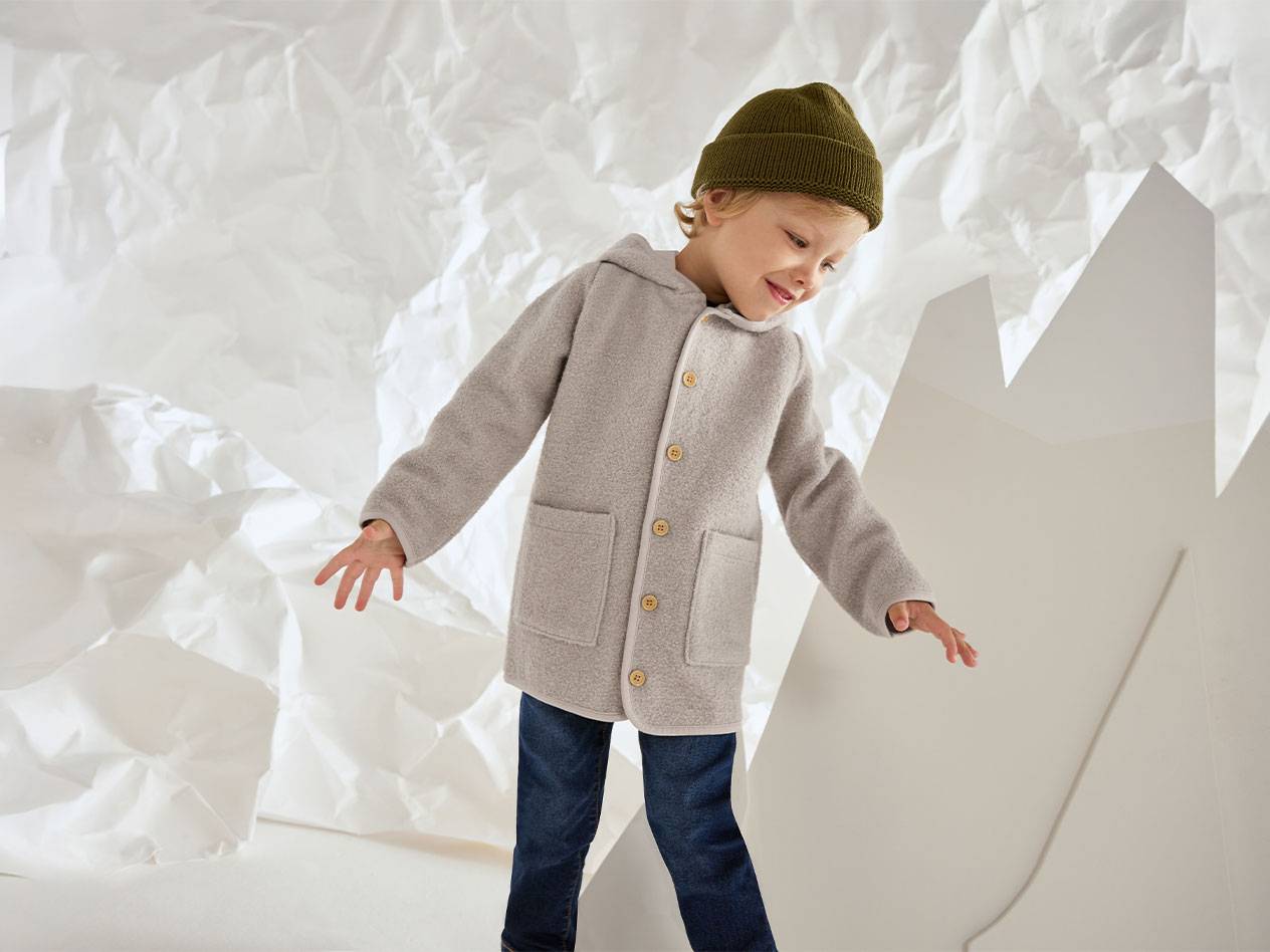 Момче със сиво вълнено яке и зелена шапка, усмихнато на фон от намачкана хартия.