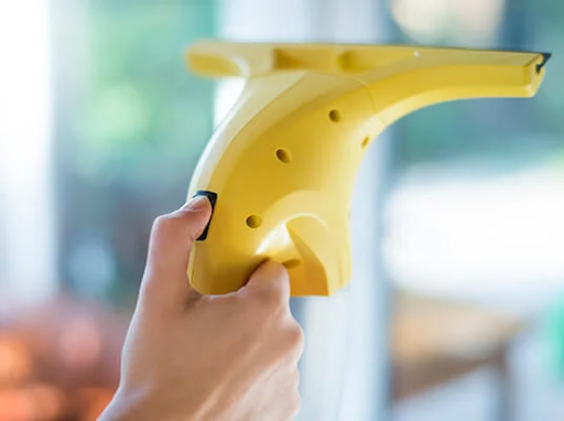 Ръка държи жълт електрически уред за почистване на прозорци, почиствайки прозорец.