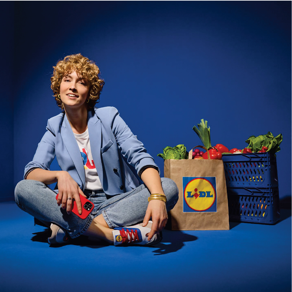 Жена седи до пазарска чанта на Lidl и щайги, пълни със свежи зеленчуци.