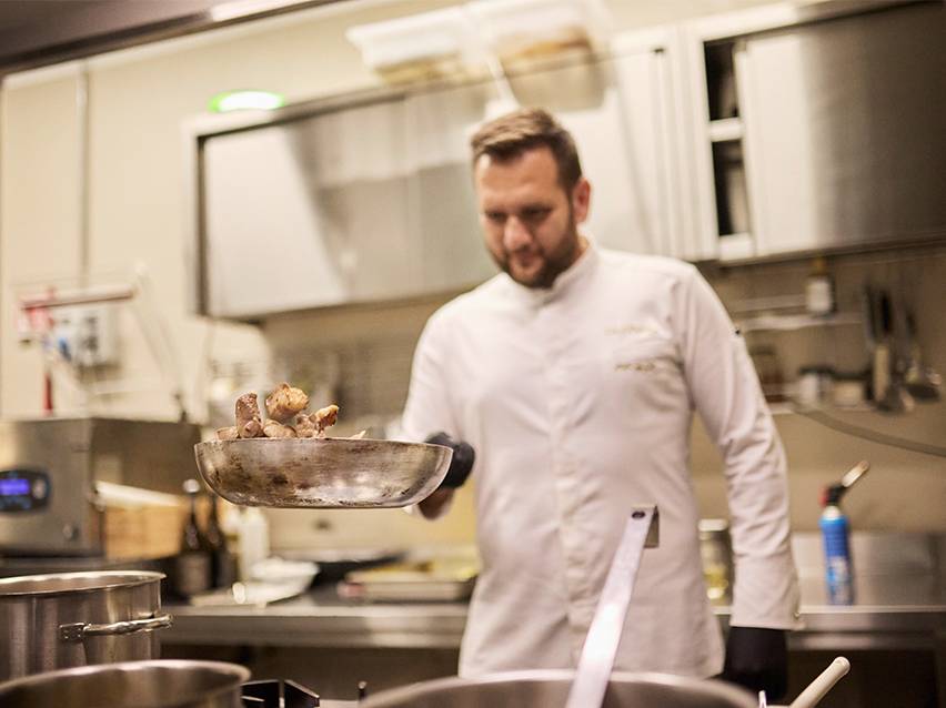 Готвач в бяла куртка хвърля храна в тиган.