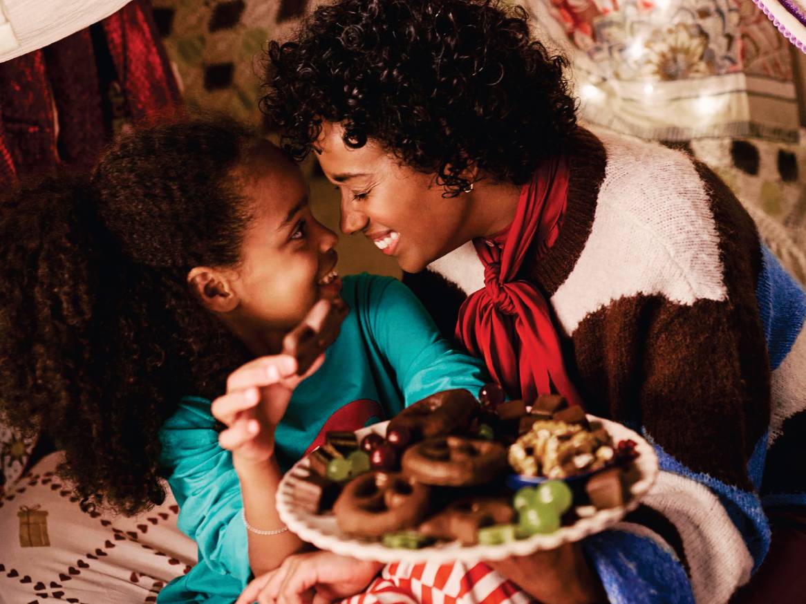 Майка и дете се усмихват, докато държат чиния с шоколадови бисквити, бонбони и плодове.