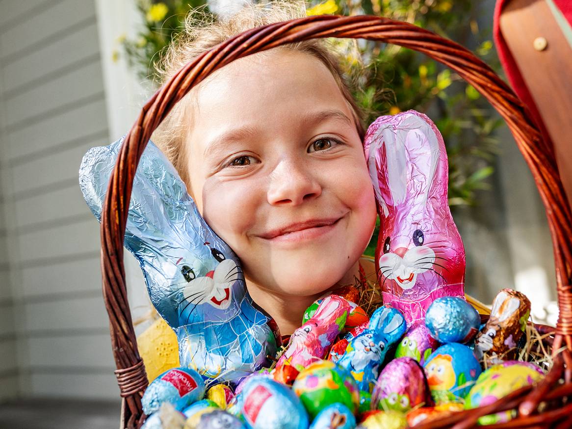Усмихнато дете с кошница, пълна с шоколадови зайчета и яйца.