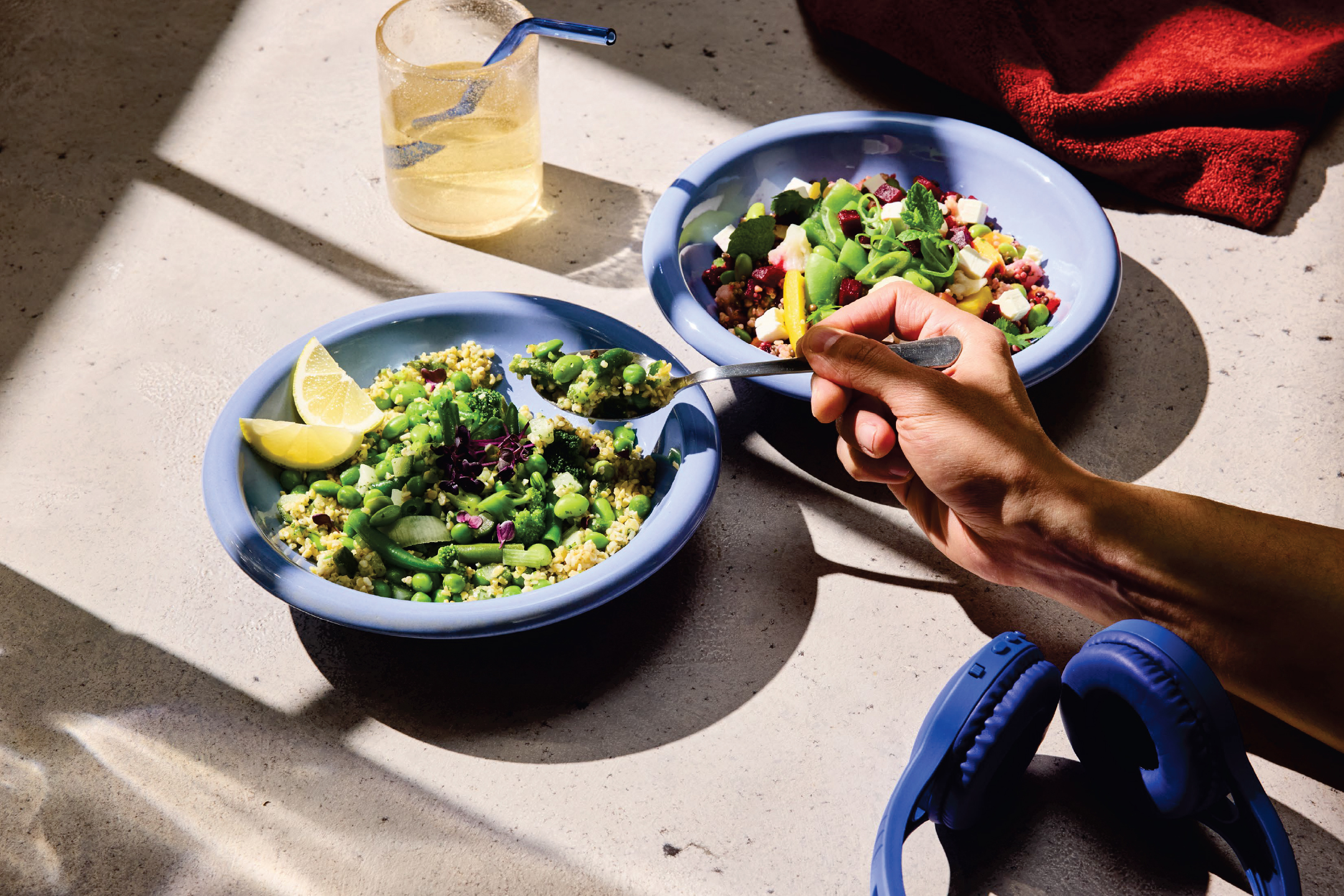 Две купи със свежи салати, напитка и слушалки на маса, осветена от слънце.
