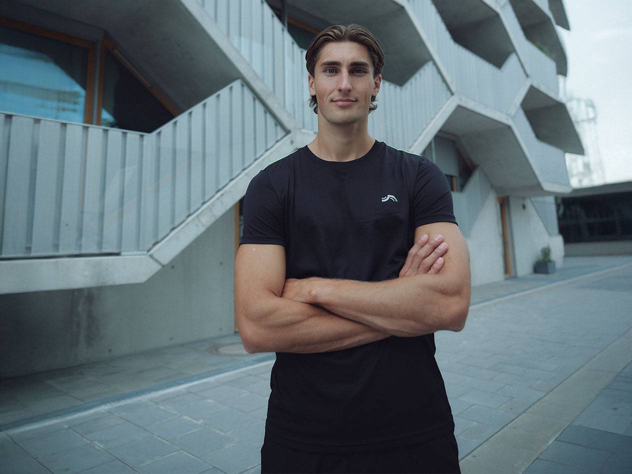 Мъж в черна тениска с лого и черни шорти, стои на открито със скръстени ръце.