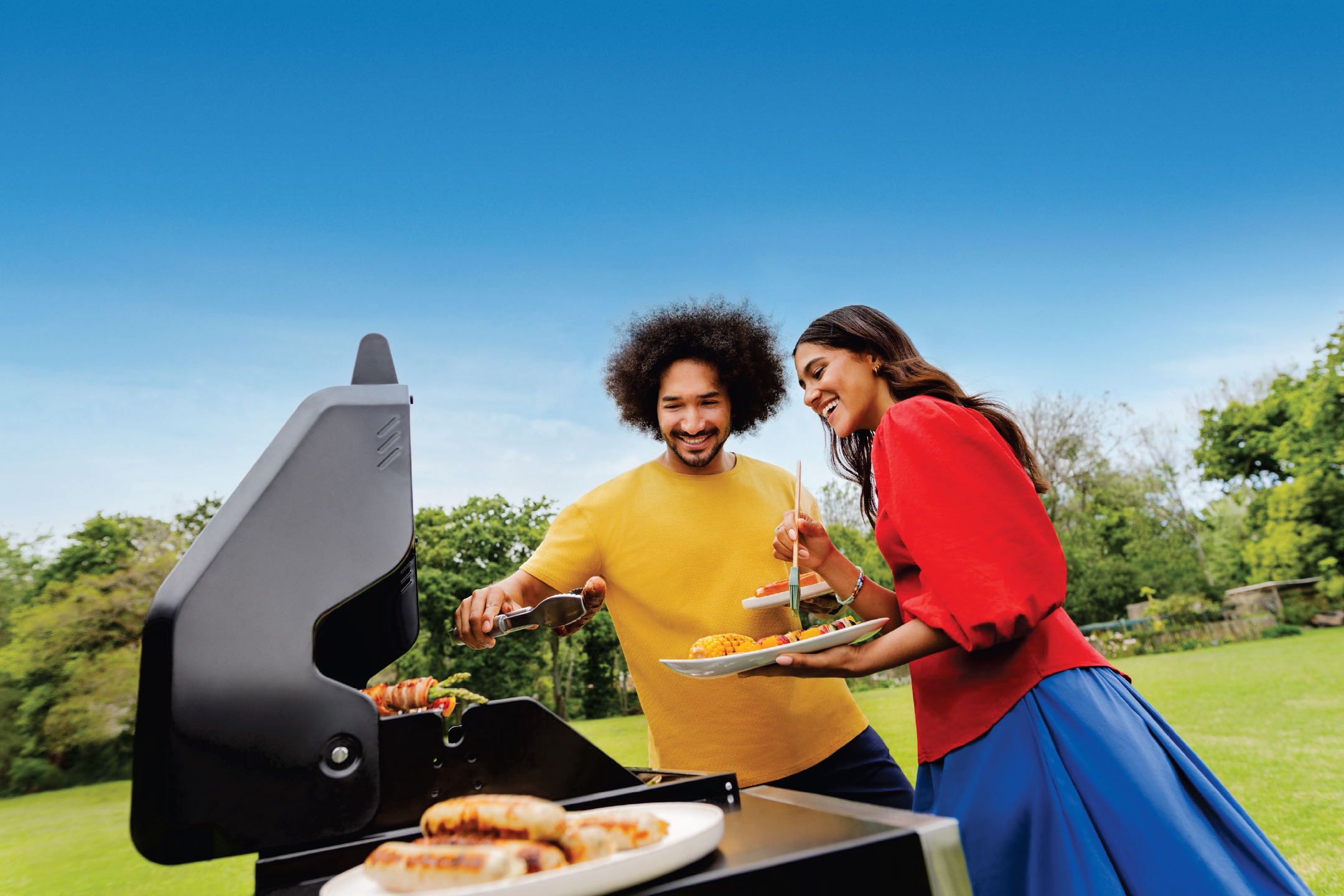 Мъж и жена пекат на скара в градината, приготвяйки наденички и зеленчуци.