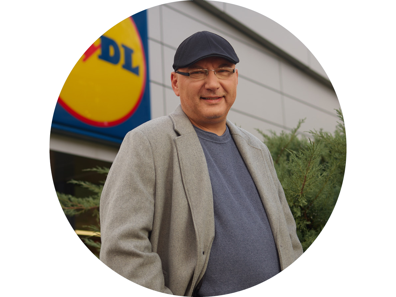 Мъж с шапка и палто пред магазин на Lidl, усмихнат.