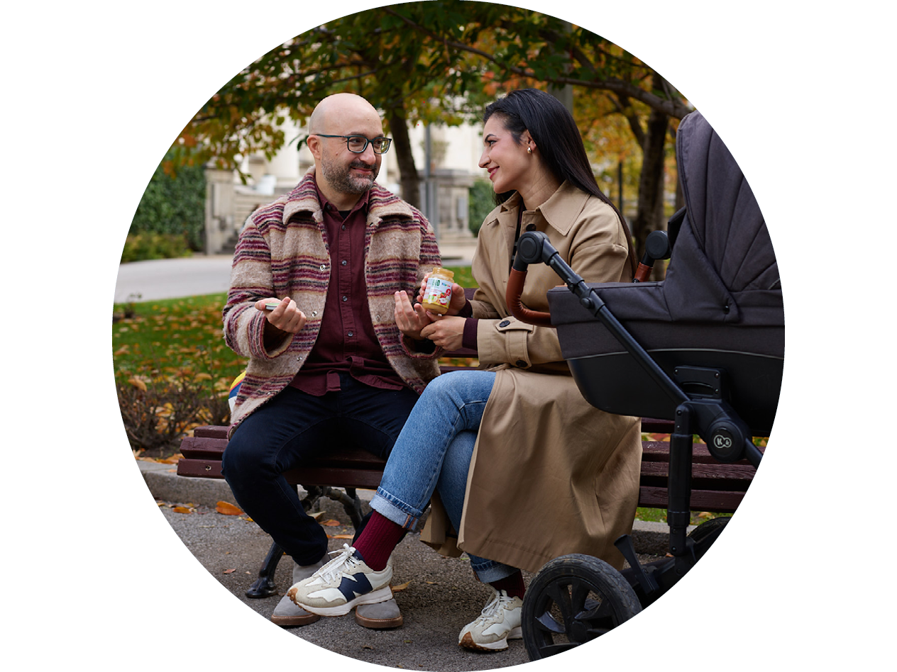 Мъж и жена на пейка в парка, хранещи бебе от бурканче с пюре до количка.