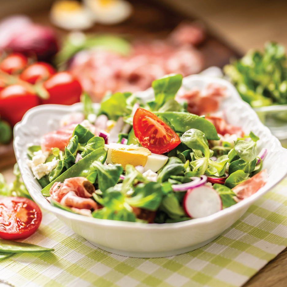 Салата с маруля, домати, яйца, шунка, сирене и репички в бяла купа на карирана покривка.