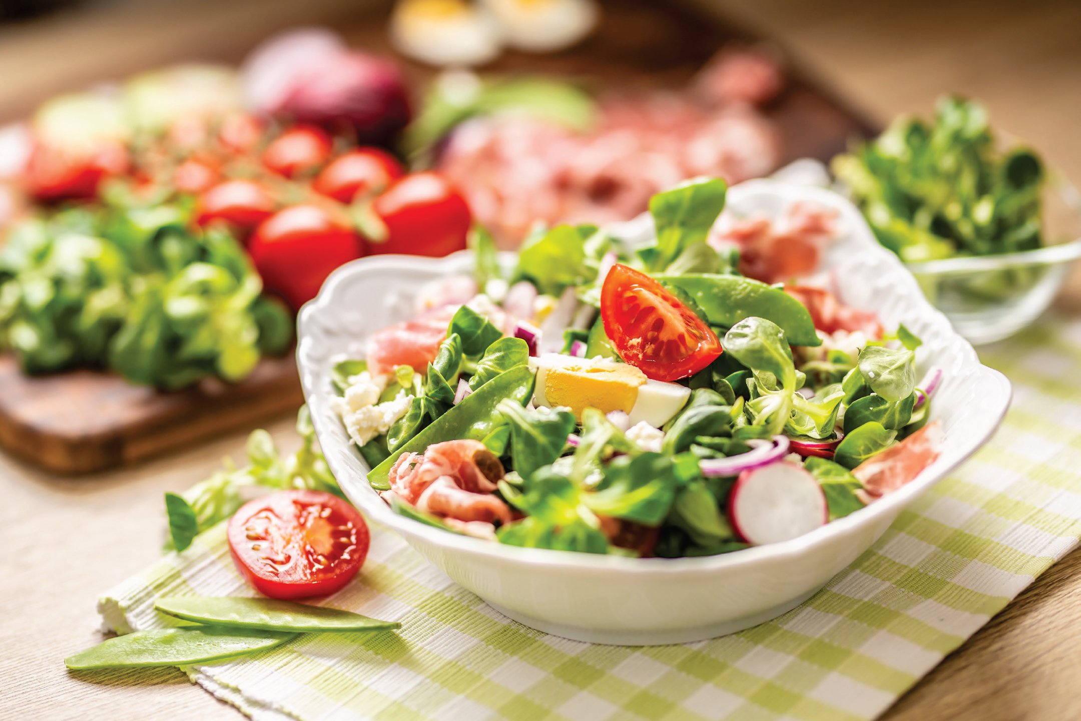 Свежа салата с маруля, домати, шунка, яйца и репички върху карирана покривка.