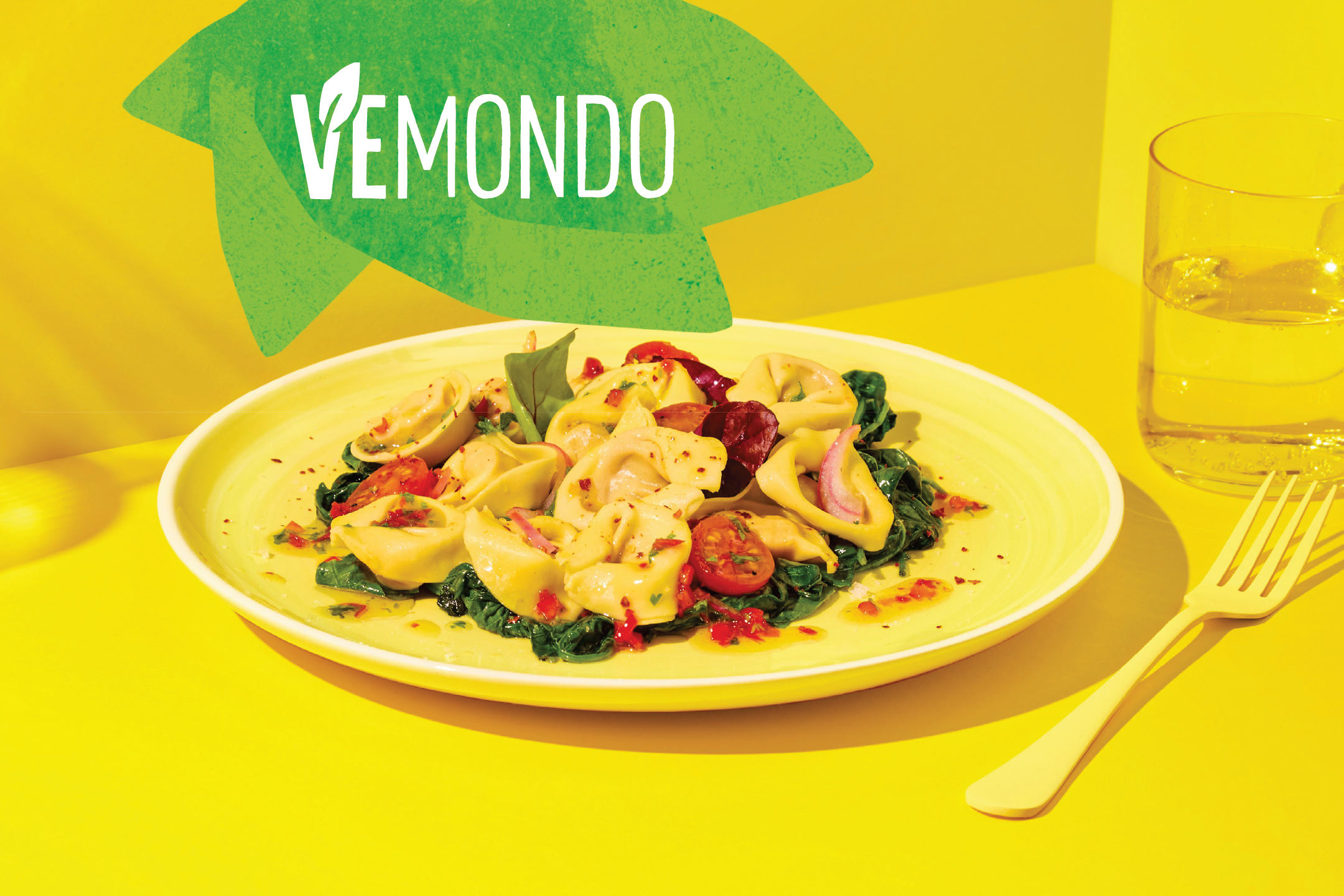 Чиния с тортелини, спанак и домати на жълт фон с чаша вода и вилица.