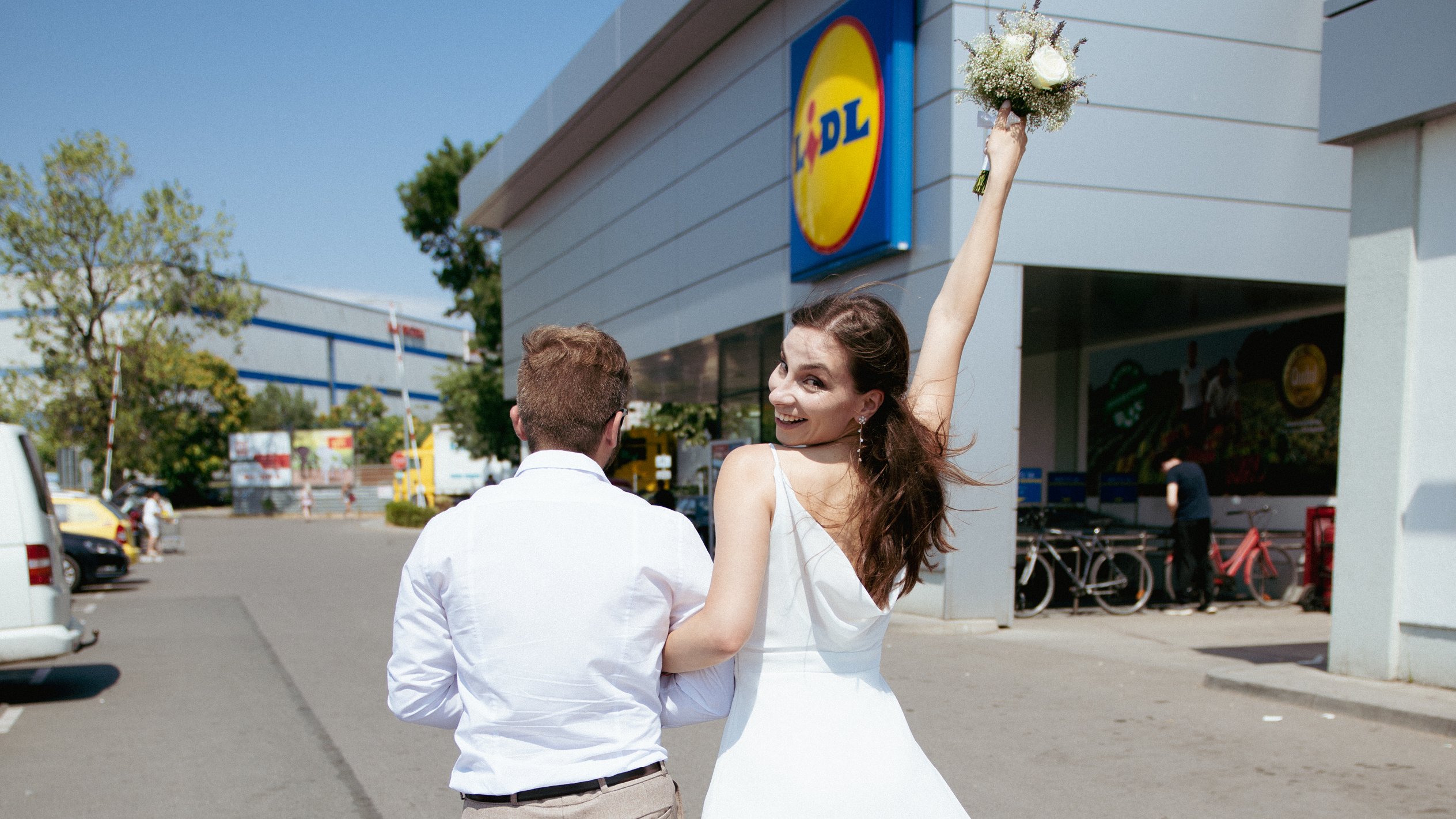 Младоженци пред магазин на Lidl, булката държи букет и се усмихва.