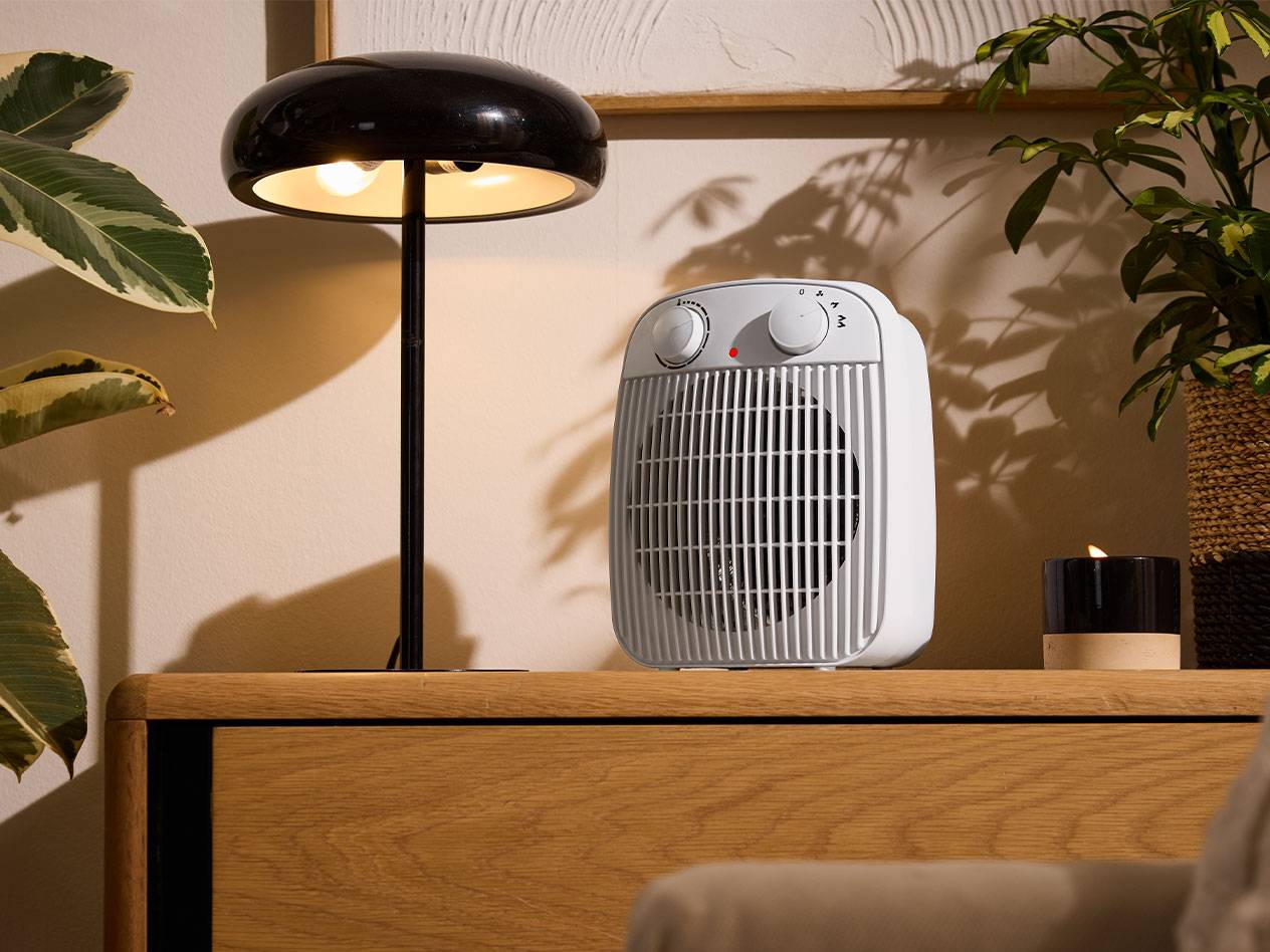 Бял нагревател и черна настолна лампа върху дървена мебел в уютна стая.