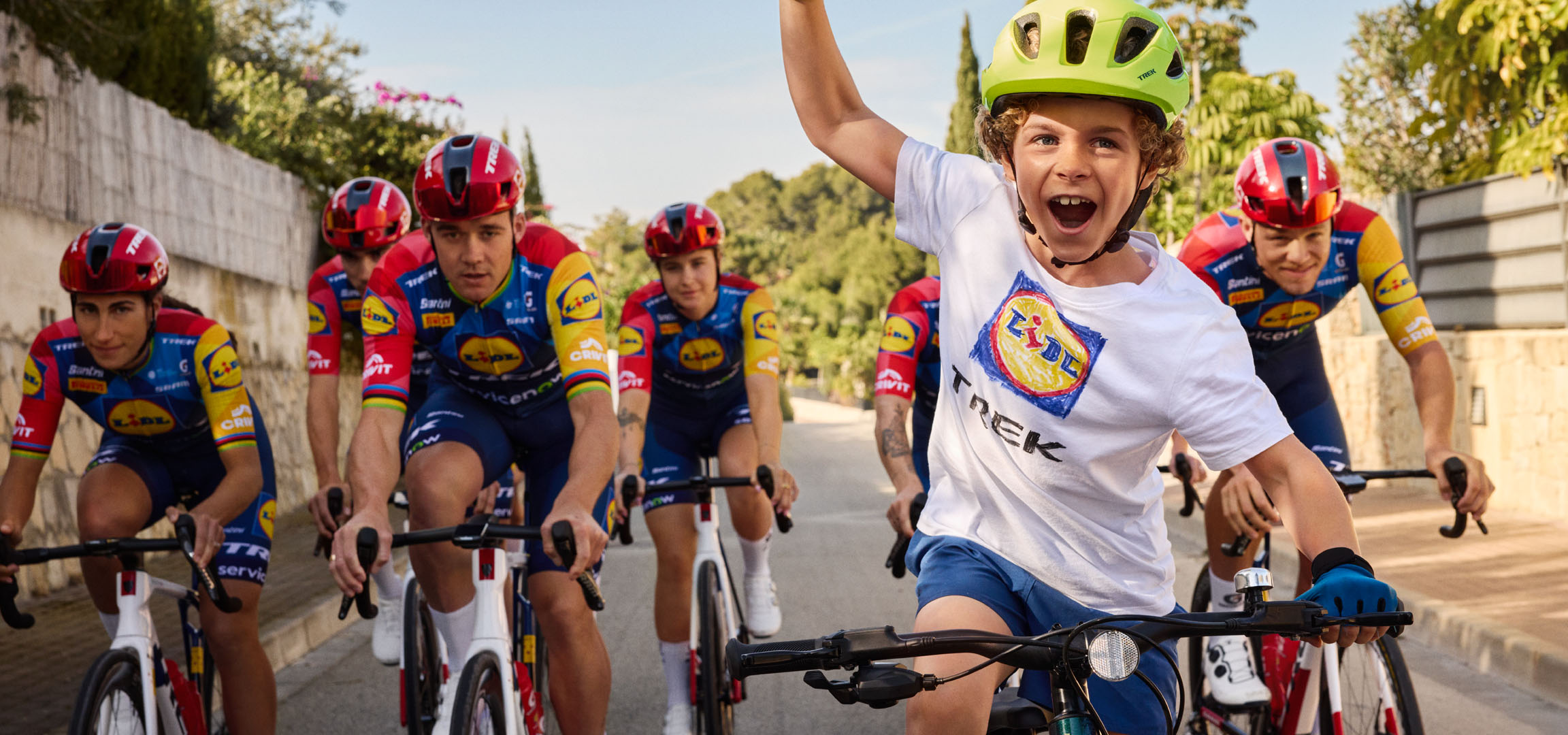 Дете с каска и тениска с логото на Lidl, заобиколено от колоездачи.