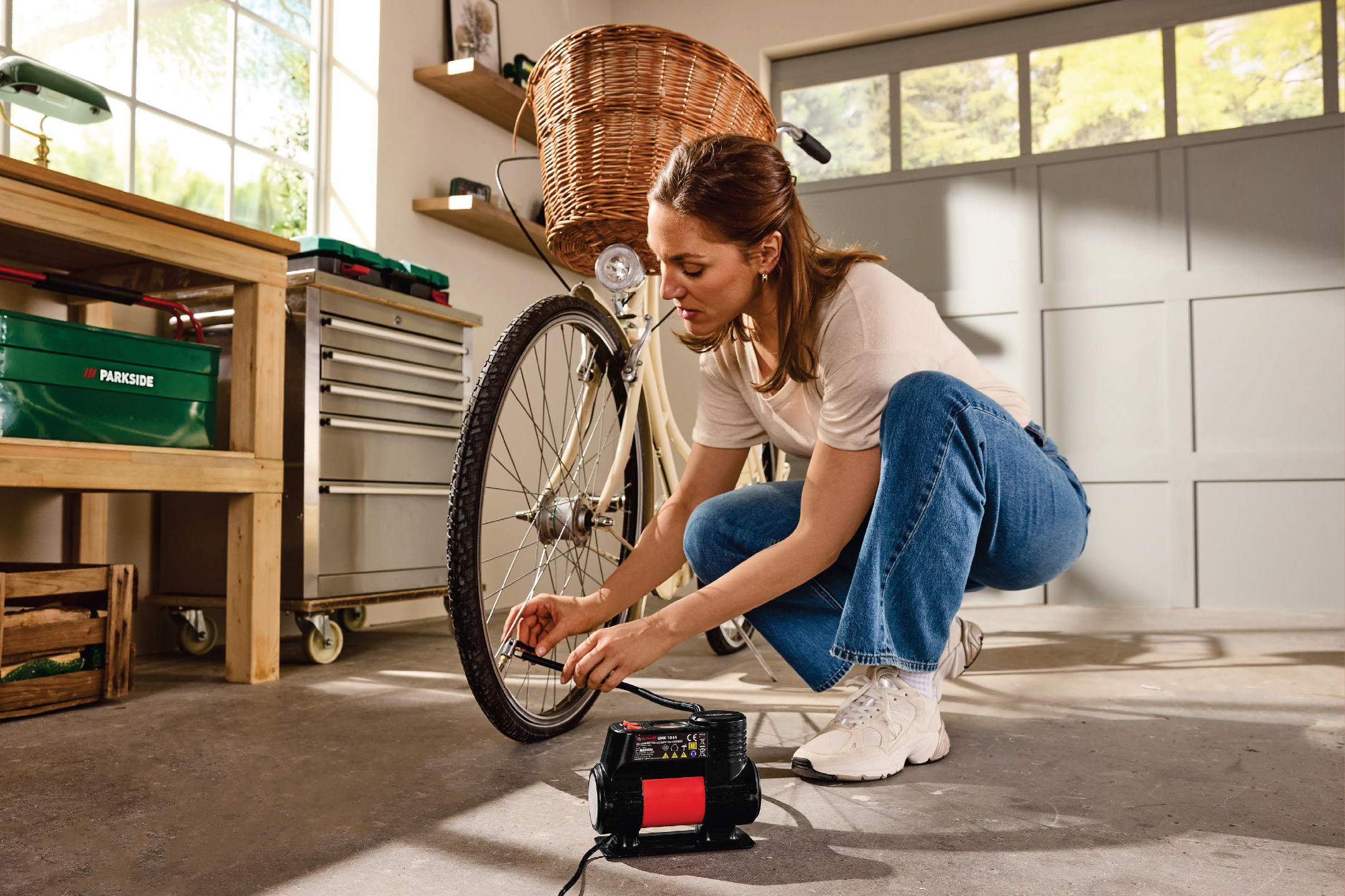 Жена помпа гума на велосипед с компресор в гараж, пълен с инструменти и рафтове.