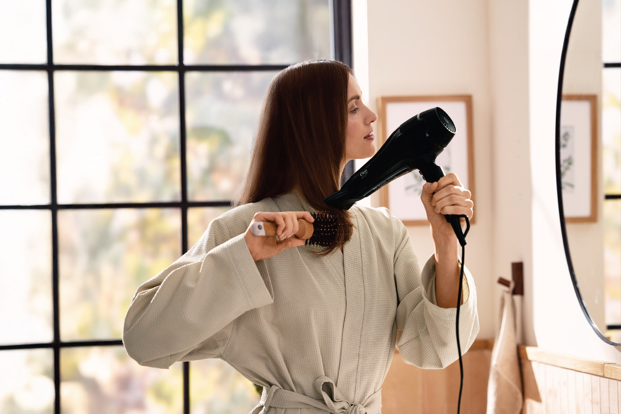 Жена в халат суши косата си със сешоар и четка за коса пред прозорец.