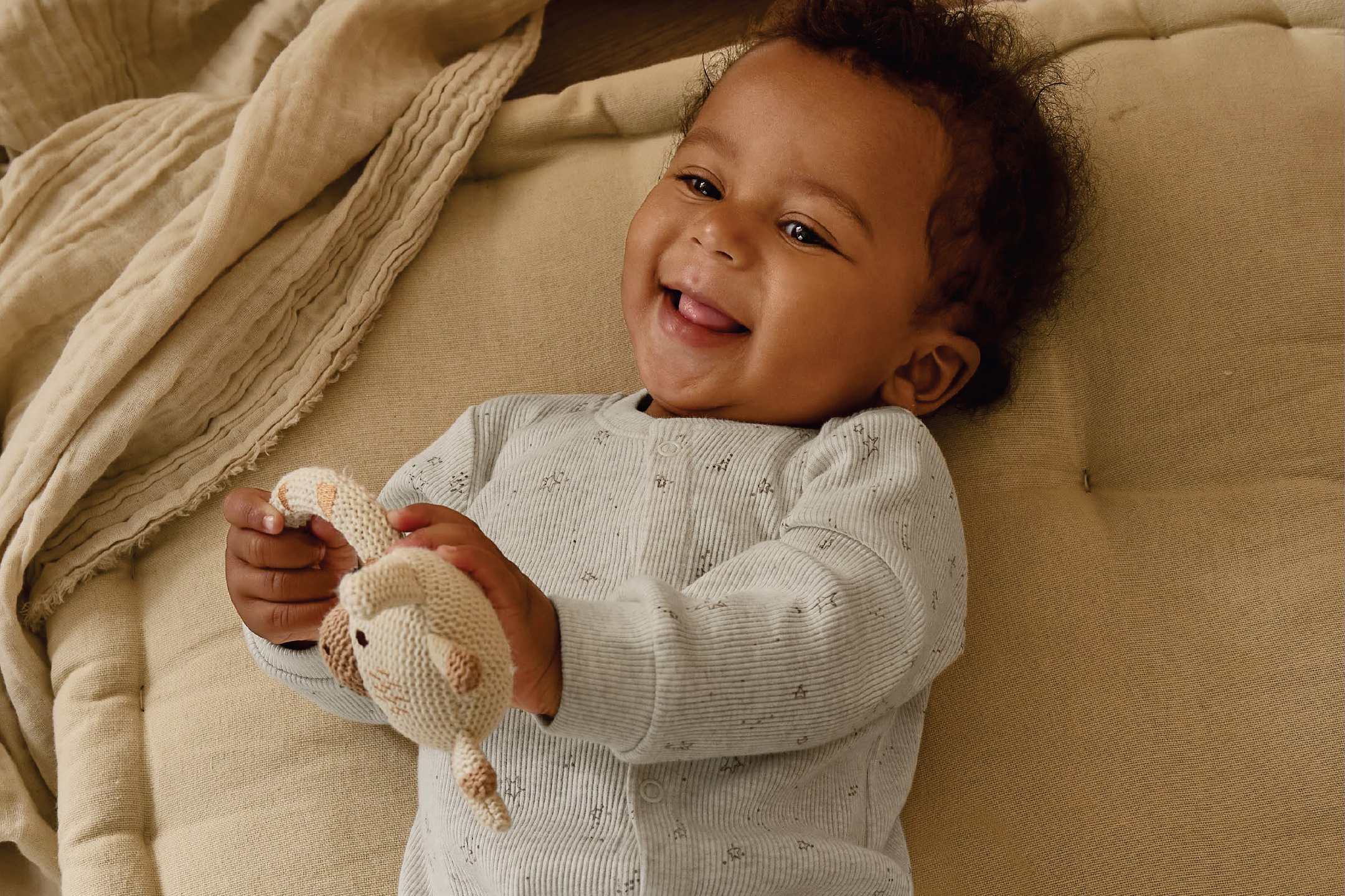 Усмихнато бебе в гащеризон, държащо плетена играчка, лежи на бежова завивка.