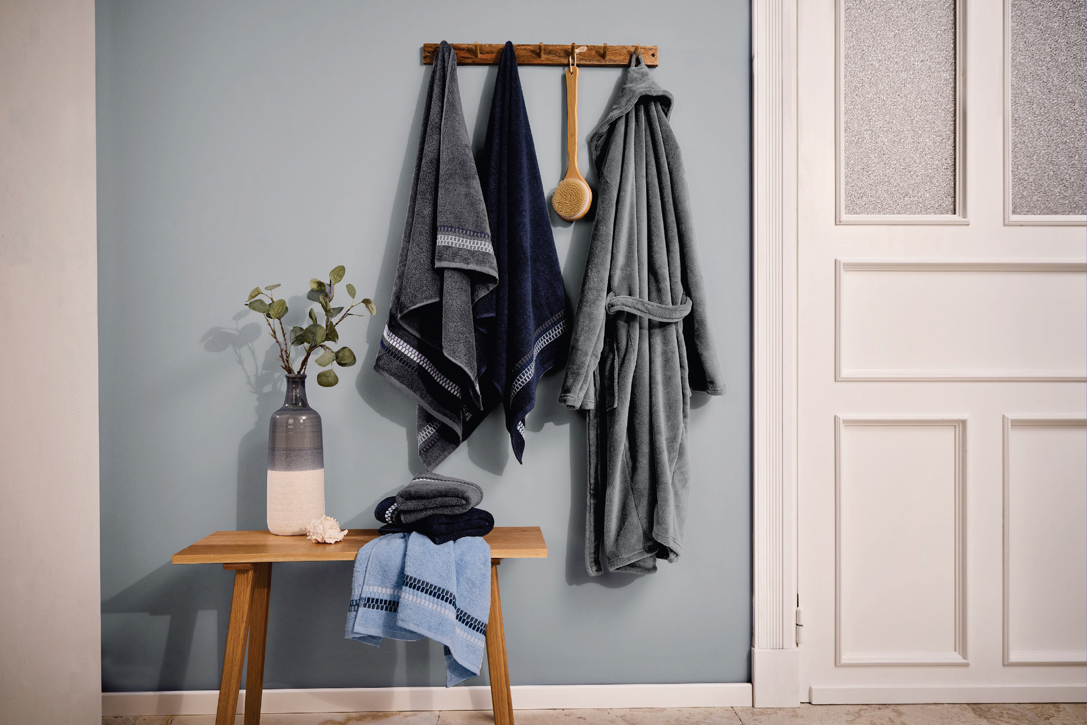Хавлиени кърпи и халат в сини и сиви нюанси, закачени на стена с дървена закачалка.