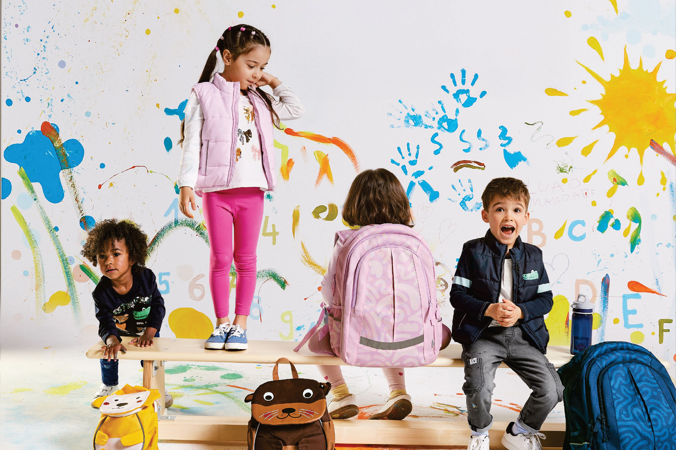 Деца с раници и жилетки на Lidl, играещи пред цветна стена.