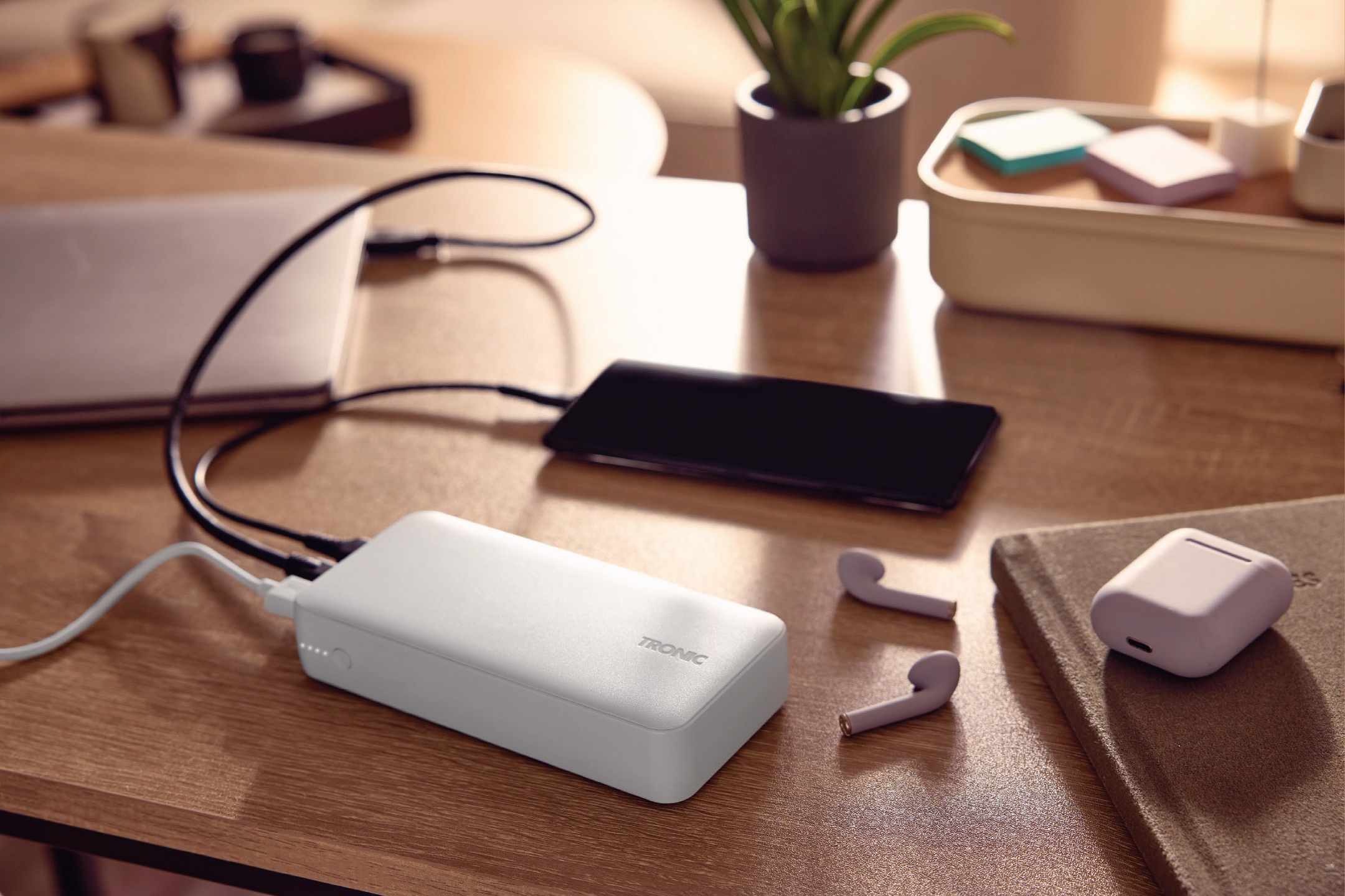 Бяла външна батерия, зареждаща телефон и лаптоп на дървена маса, с безжични слушалки и кутия за съхранение.