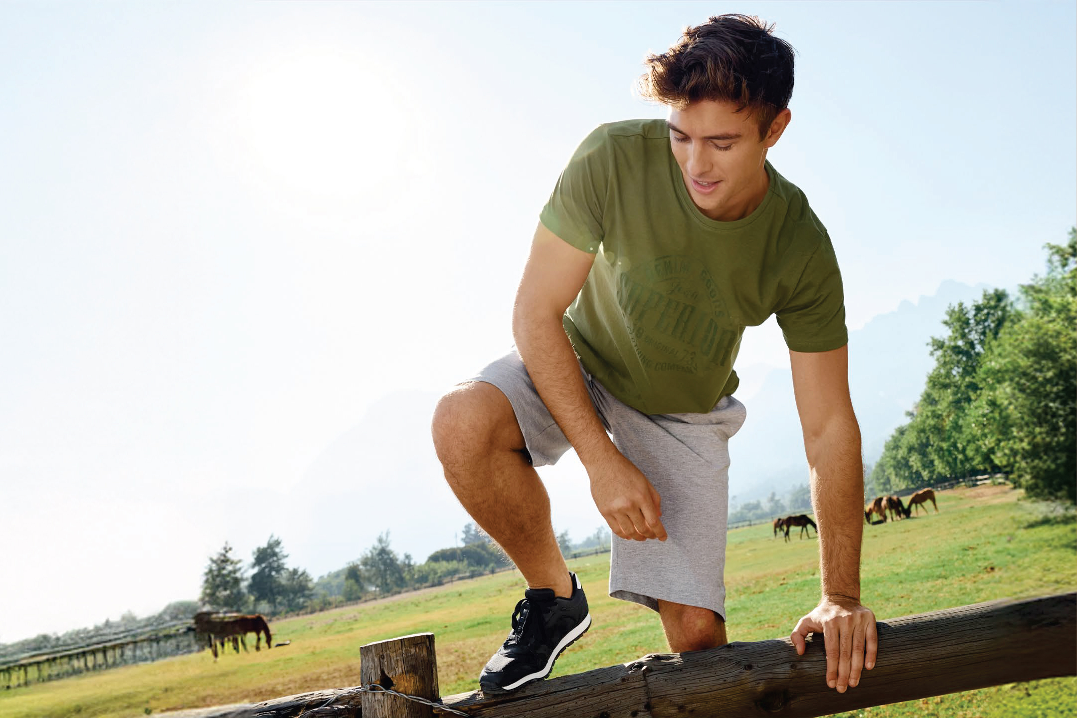 Мъж в зелена тениска и сиви къси панталони се катери по дървена ограда на поле с коне.