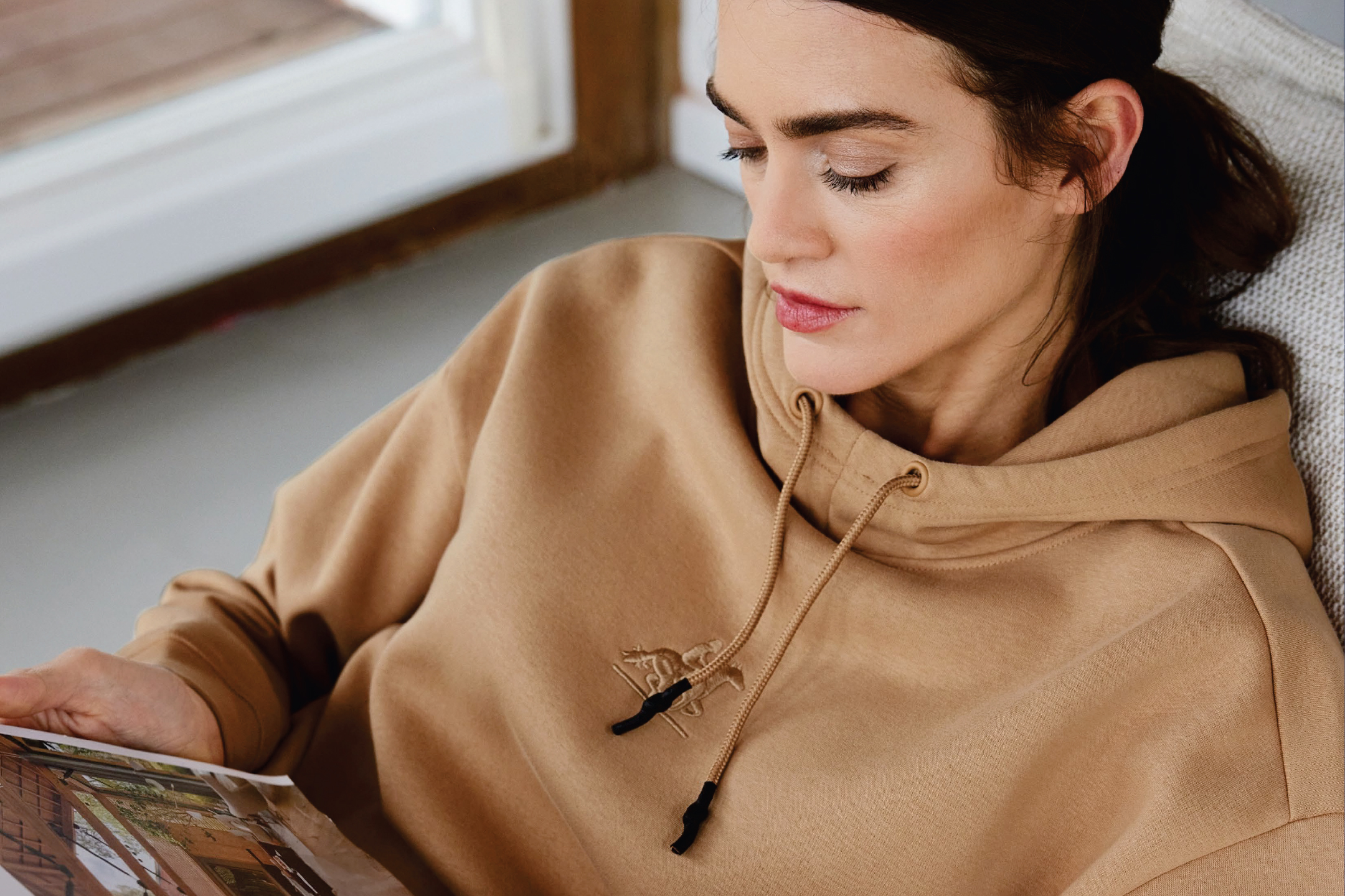 Жена в бежов суичър с качулка чете списание, отпуснато в кресло.