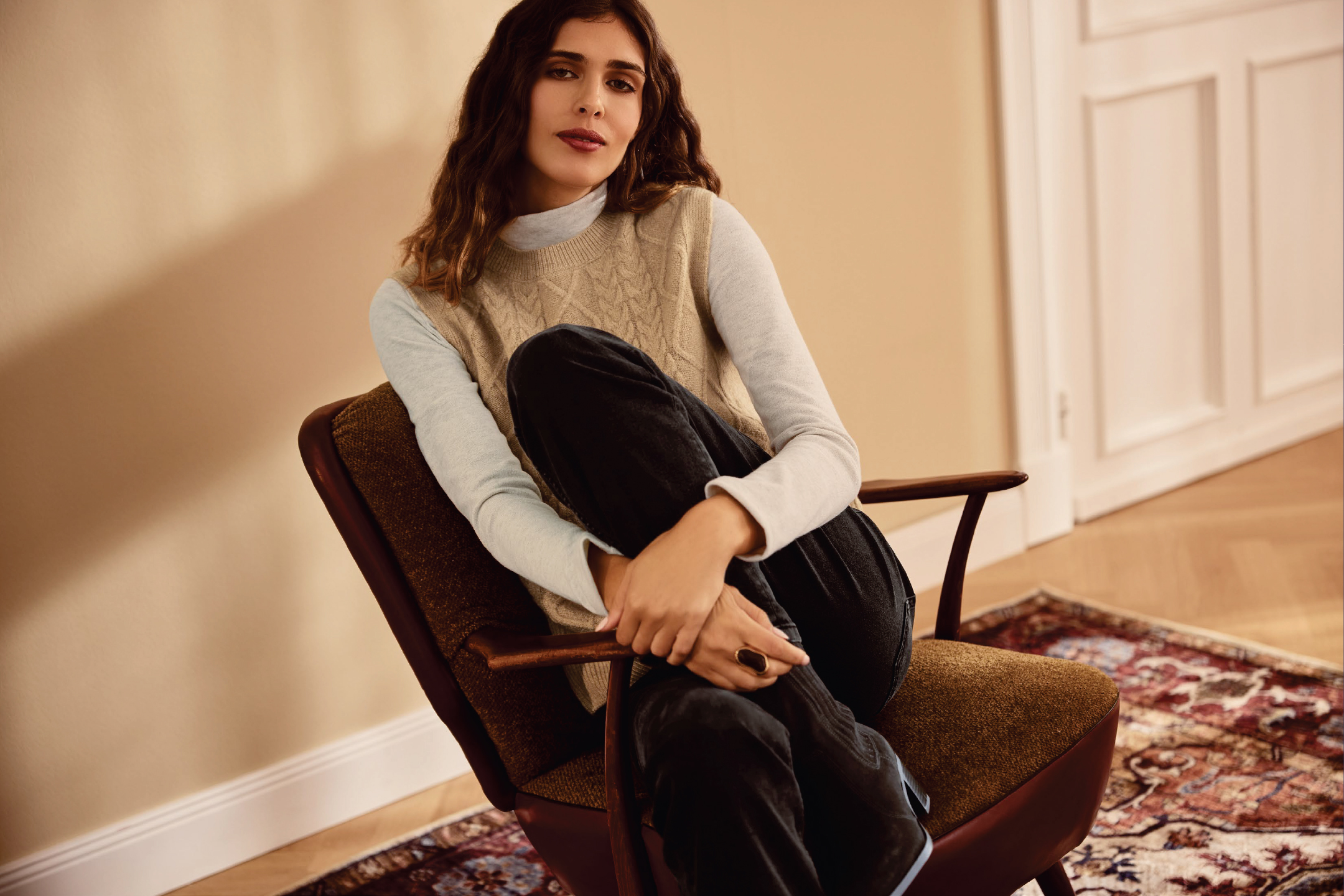 Жена в плетен елек и панталон, седнала на стол в стая с килим.