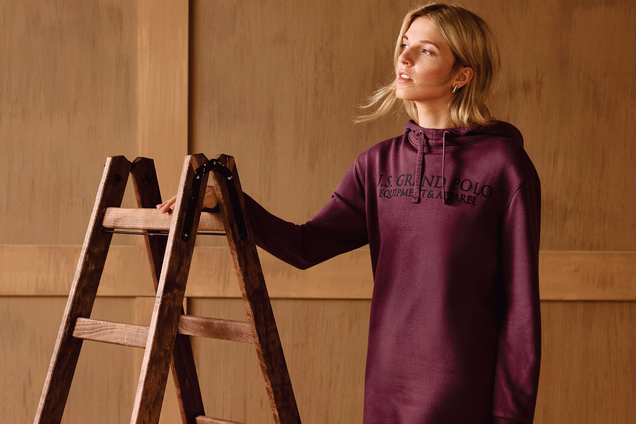 Жена в бордо суичър с качулка, облегната на дървена стълба.